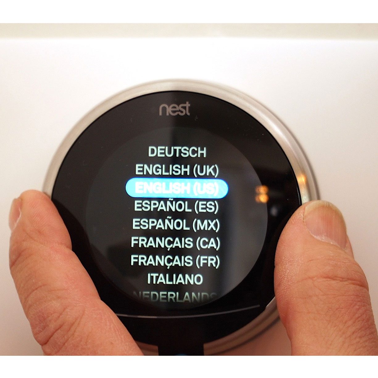 A hand interacts with a digital thermostat display, selecting "ENGLISH (US)" from a list of language options on a round screen in a home environment.