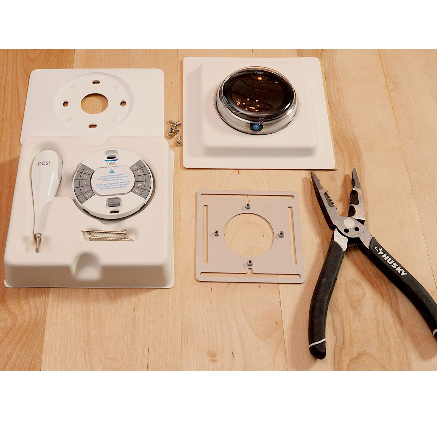 A Nest thermostat kit, including tools and mounting plates, is arranged on a wooden surface, indicating preparation for installation in a home setting.
