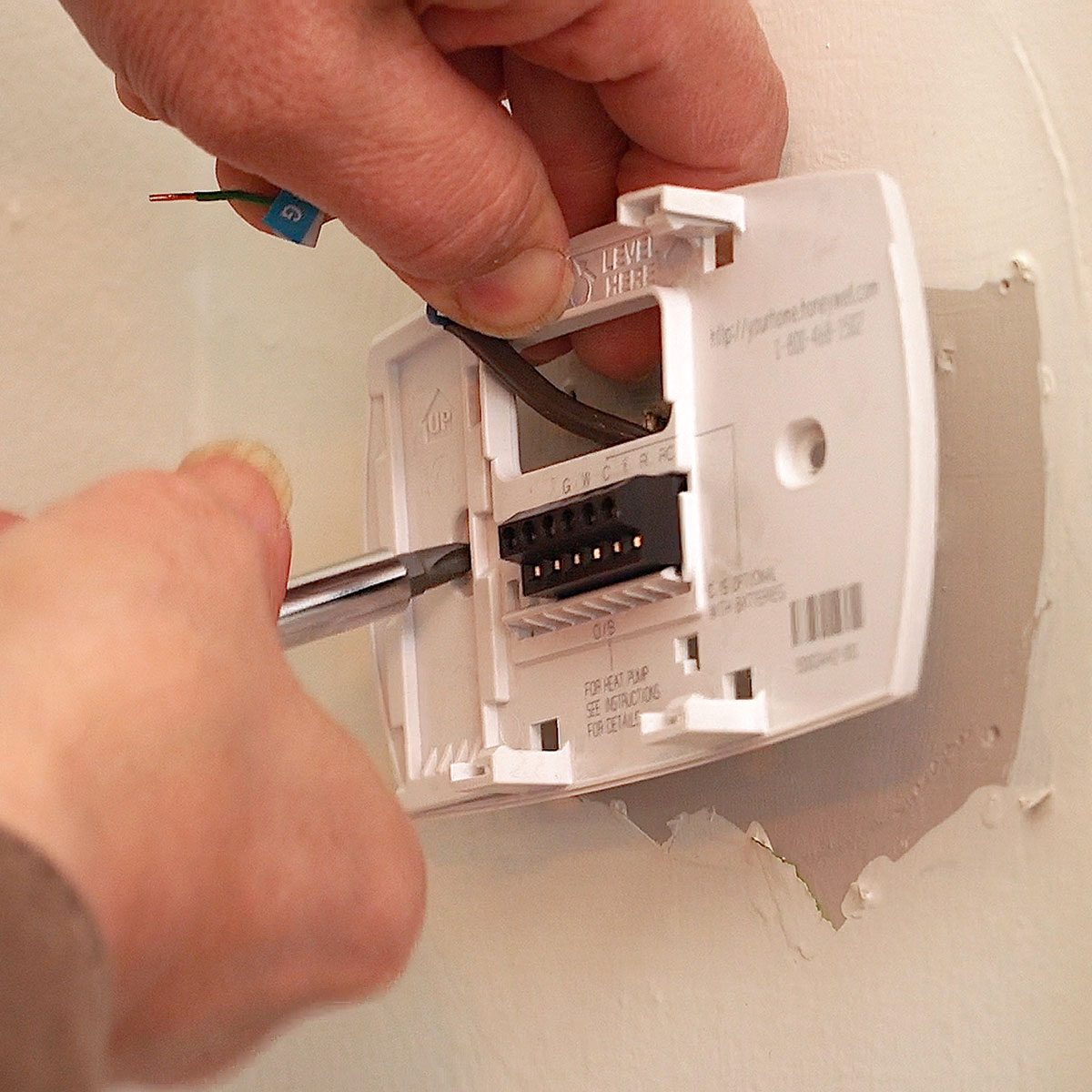 A person is installing a thermostat on a wall, using a screwdriver to secure wires inside the device. The wall is partially painted.