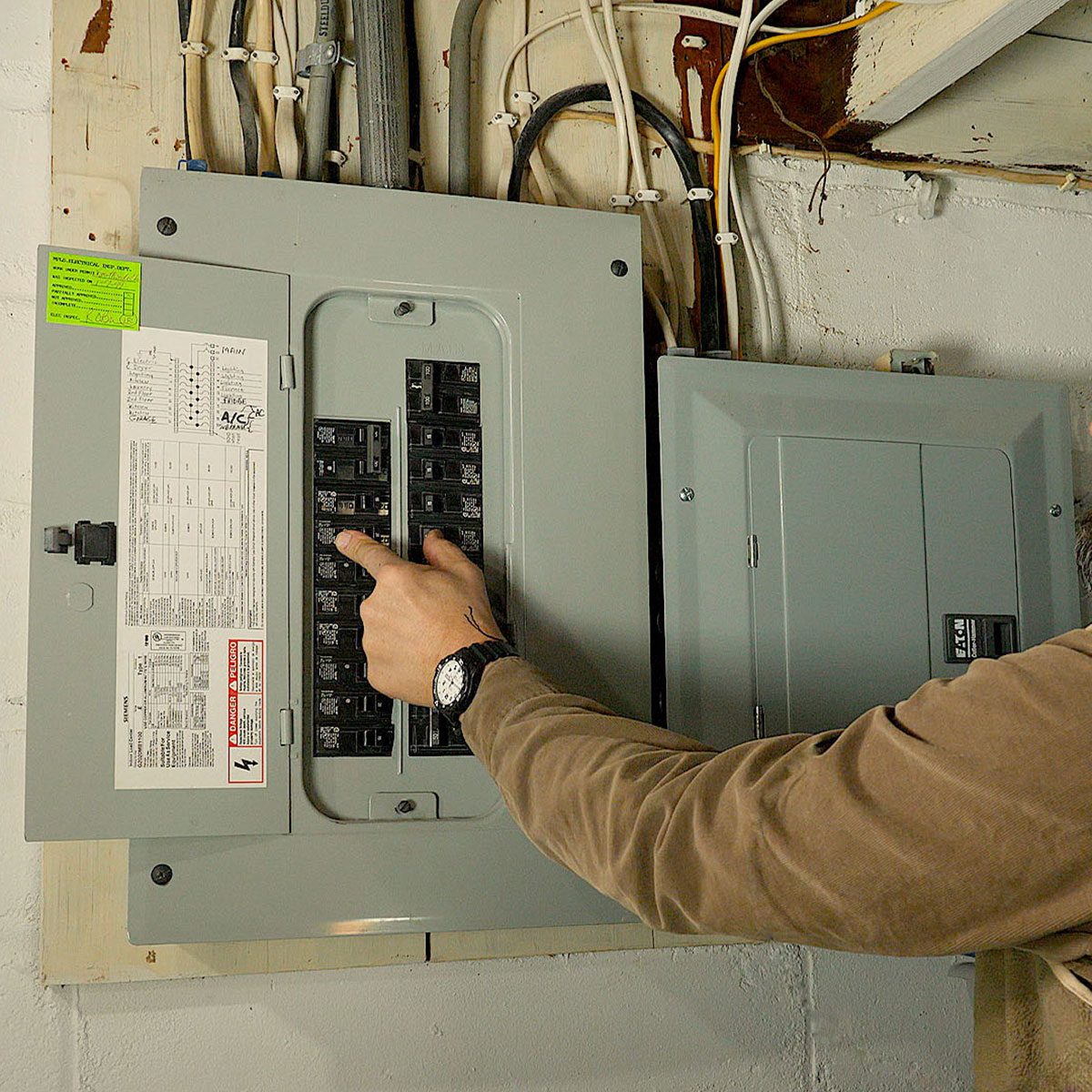 A hand points to a circuit breaker in a gray electrical panel mounted on a wall, surrounded by wires and a utility area.