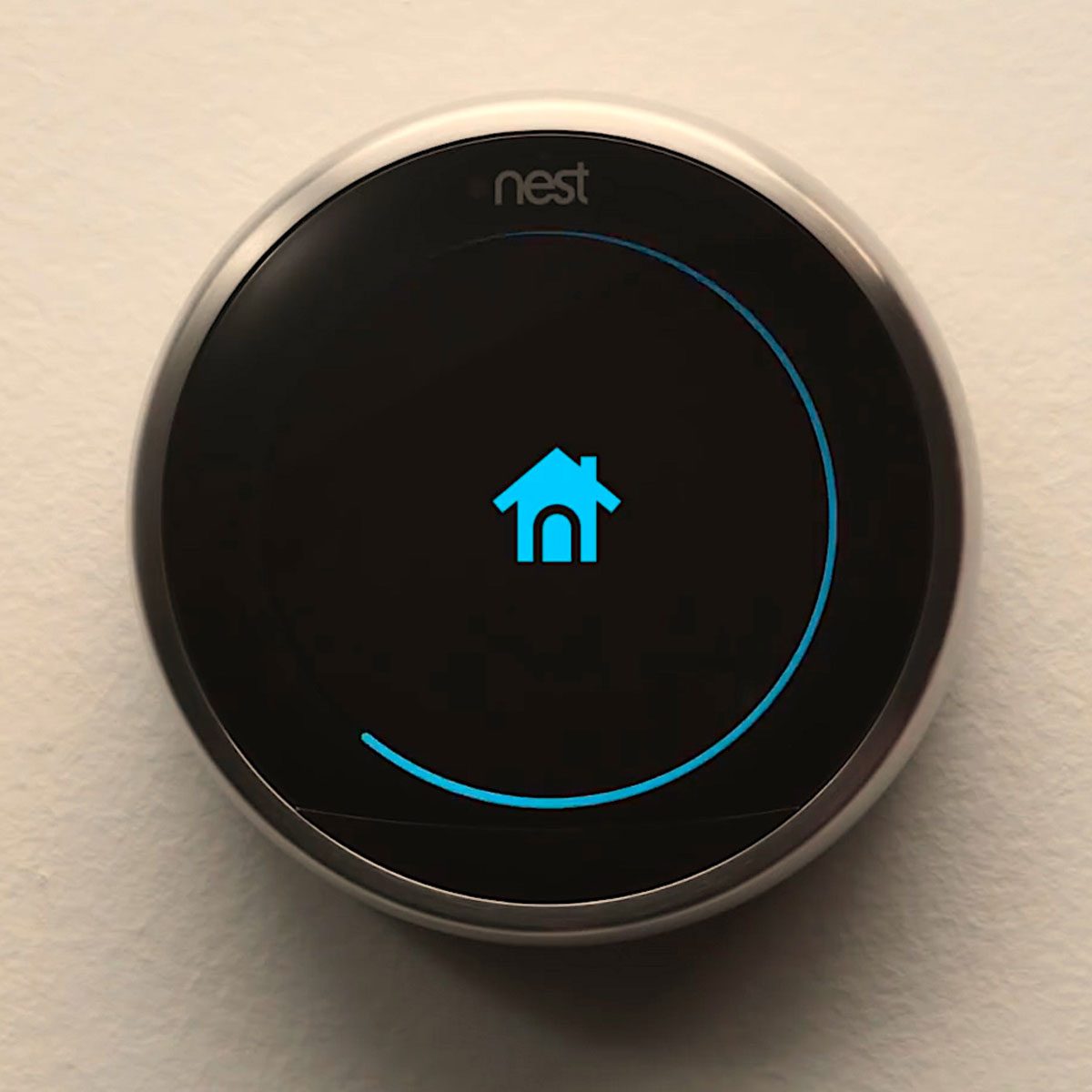 A circular digital thermostat displays a blue house icon while mounted against a light-colored wall, indicating a home setting.