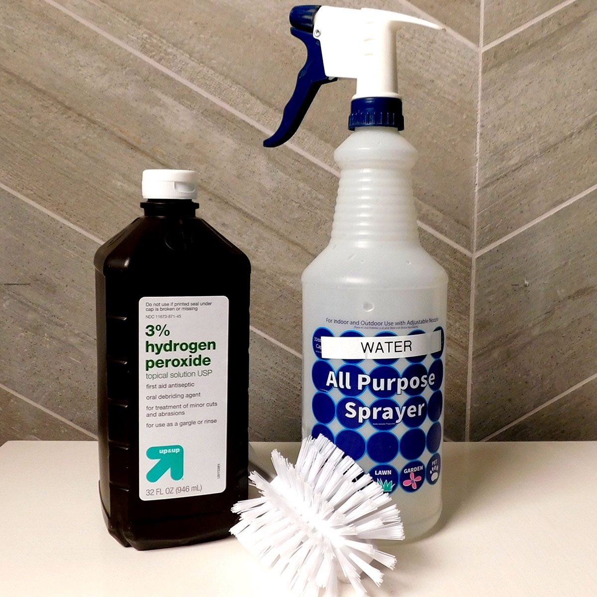 A bottle of hydrogen peroxide stands next to a spray bottle labeled "WATER All Purpose Sprayer," alongside a white cleaning brush, on a tiled surface.