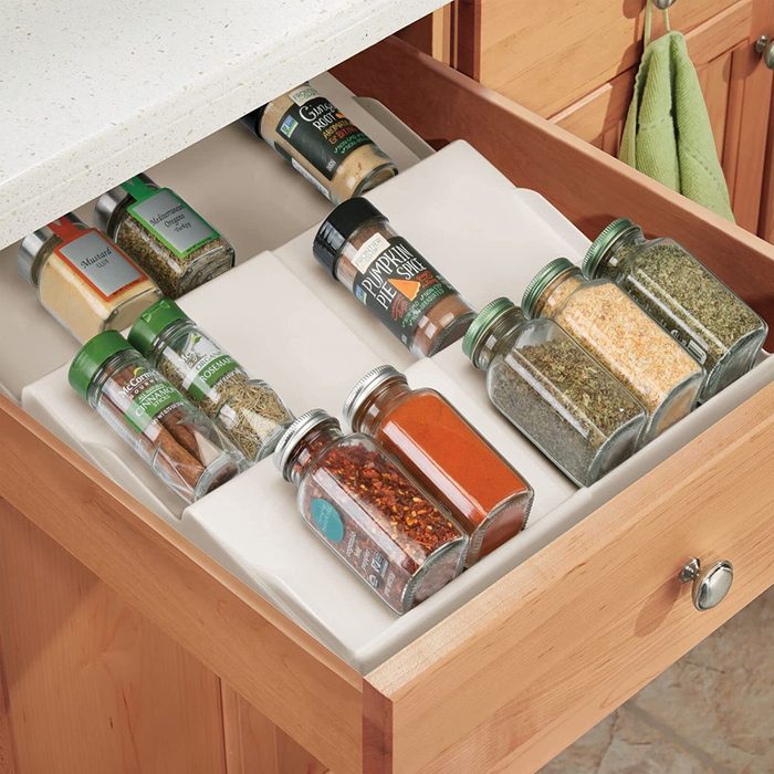 Spice jars are neatly organized in a kitchen drawer, with labels facing up, surrounded by a light-colored liner and wooden drawer sides.