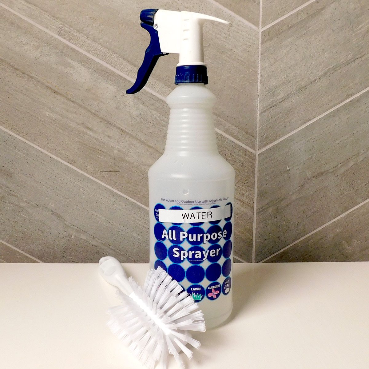 A clear bottle labeled "Water All Purpose Sprayer" sits next to a white cleaning brush against a textured tile background.