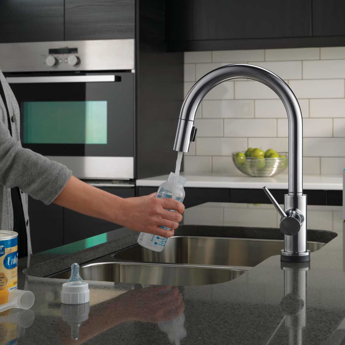 A person fills a baby bottle with water from a modern kitchen faucet, set against a sleek countertop and cabinets, with a bowl of green fruit nearby.