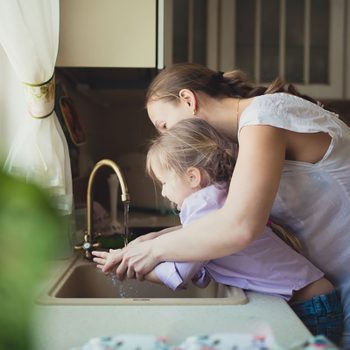 A woman helps a girl wash her hands at a kitchen sink, surrounded by sunlight and kitchen decor. Water flows from the faucet.