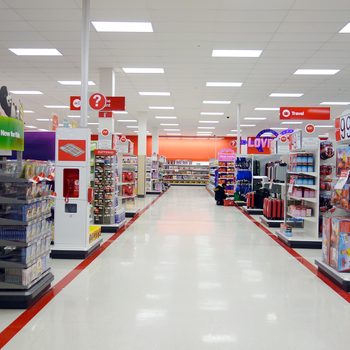 Shelves filled with various products line a brightly lit retail aisle, while clear signage directs shoppers towards different departments like travel and batteries.