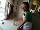 A person in a green shirt and apron mixes batter in a bowl on a table, with sunlight illuminating a window and curtains in the background.