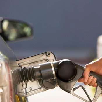 A fuel nozzle is being held and inserted into a car's fuel filler opening at a gas station, with a blurred background of a vehicle.