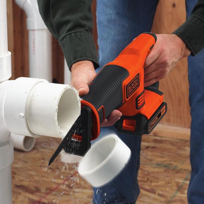A person is using a cordless saw to cut a white PVC pipe in a workshop, causing debris to fall from the cut section.