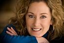 A woman with curly hair smiles warmly while resting her arms on a surface, set against a softly blurred background.