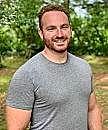A smiling man stands in a green outdoor setting, wearing a gray t-shirt, with trees and foliage in the background.
