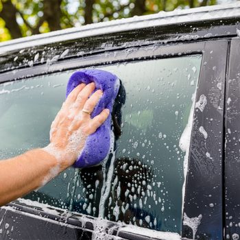 A hand holds a soapy purple sponge, washing a car window. Bubbles and water droplets surround the glass in a bright outdoor setting.