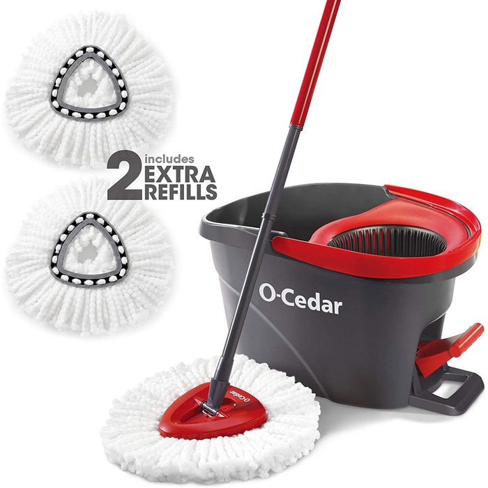 A mop with a triangular head and a red handle is leaning against a gray bucket, accompanied by two extra white mop refills.