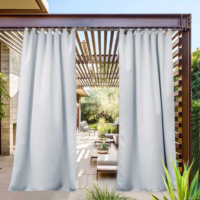Light gray curtains hang from a wooden pergola, framing a sunny patio with modern furniture and greenery, creating an inviting outdoor space.