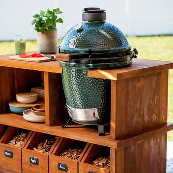 A green ceramic grill rests on a wooden shelf, surrounded by food preparation items and storage boxes filled with different types of wood chips outdoors.