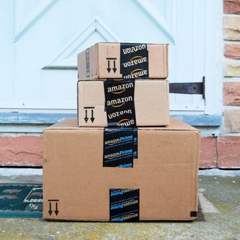 Three stacked cardboard boxes with Amazon branding sit on a doorstep, surrounded by a stone facade and a decorative doormat.