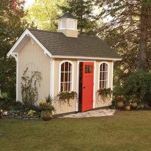 A quaint garden shed with a red door stands on a stone path, surrounded by vibrant flowers and greenery under a clear sky.