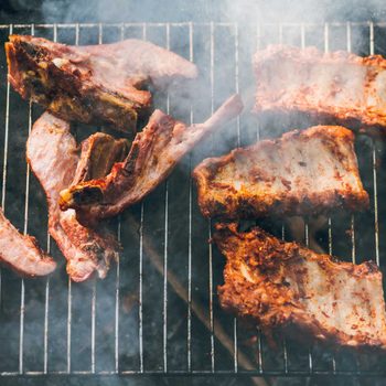 Meat ribs are grilling on a smoky barbecue rack, visibly seared and coated in spices, surrounded by a haze of smoke.