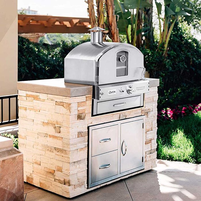 A stainless steel pizza oven sits on a stone base, prominently placed in a sunny, outdoor patio surrounded by greenery and colorful flowers.