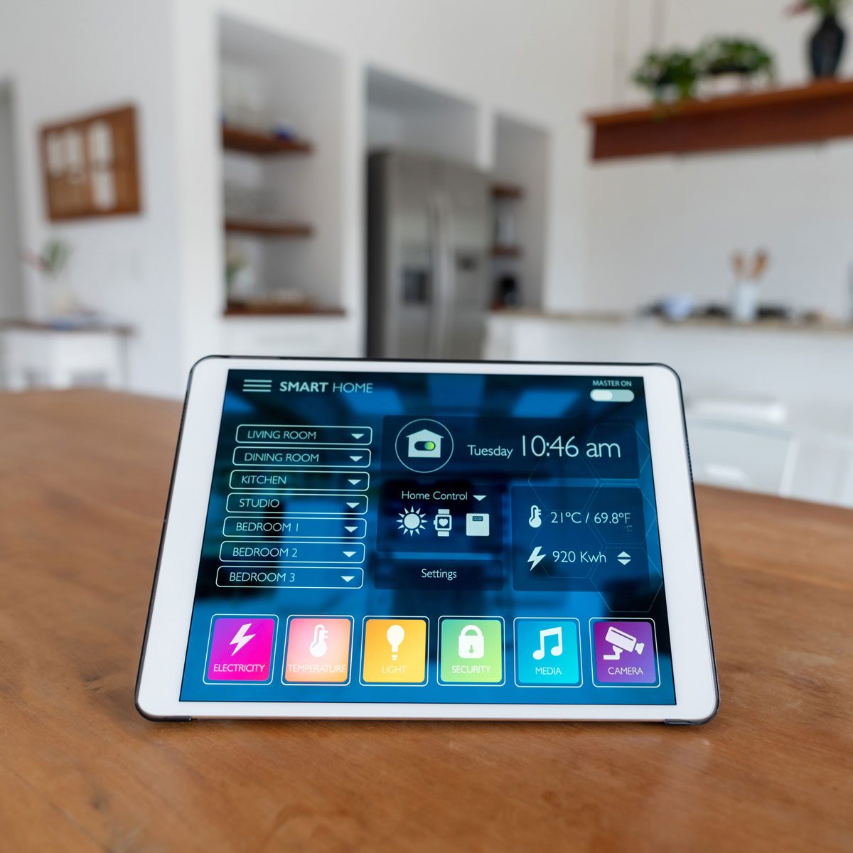 A tablet displays smart home controls, showing room statuses and settings in a modern kitchen environment with wooden shelves and appliances in the background.