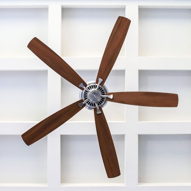 A ceiling fan with wooden blades spins slowly, suspended from a white grid-patterned ceiling, providing a cooling breeze in a bright room.