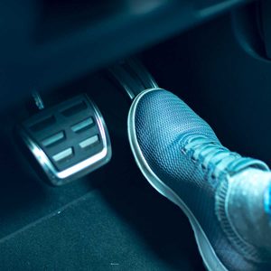 A blue sneaker presses down on a car pedal, indicating acceleration, within a dimly lit vehicle interior.