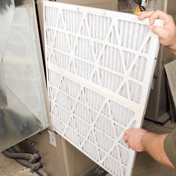 A person is removing an air filter from a furnace, surrounded by machinery and ducts in a maintenance environment.