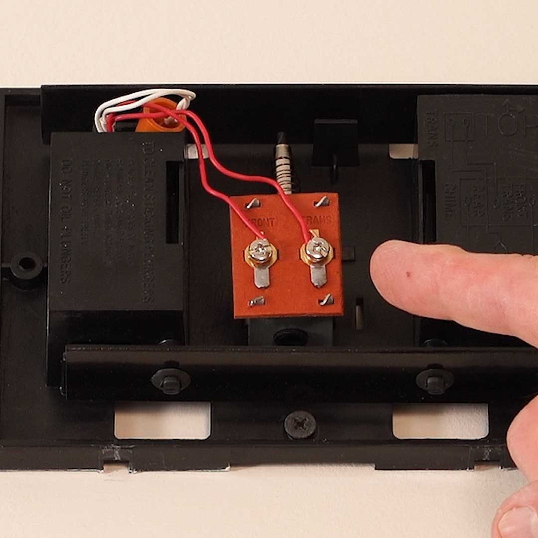A person's finger touches a circuit board inside a device, with wires and components visible against a neutral background, indicating examination or repair.