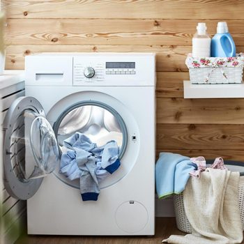A washing machine is open, filled with clothes. It sits in a wooden-paneled room alongside laundry products and a basket with additional laundry items.