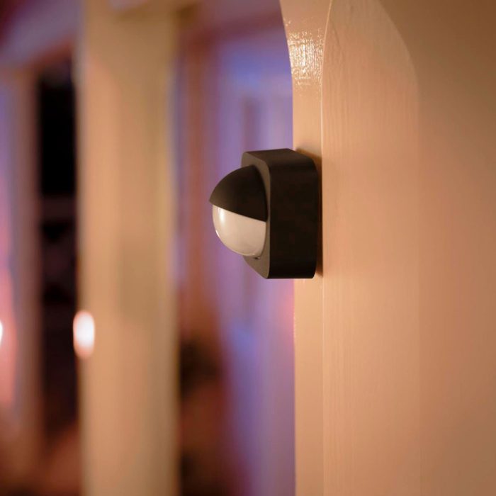 A black motion sensor light is mounted on a wall, illuminating a softly lit corridor with warm tones and shadows in the background.