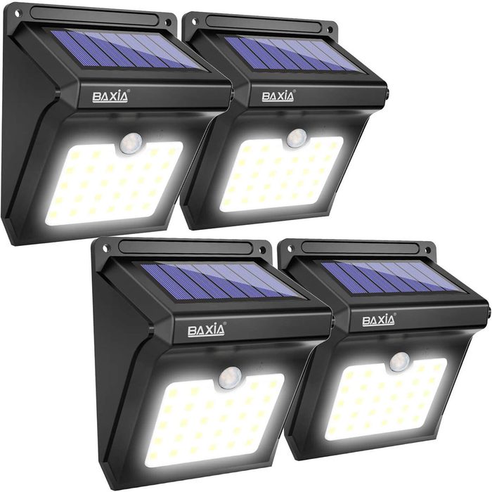 Four black solar-powered lights with motion sensors illuminate brightly, designed for outdoor use, featuring solar panels at the top for energy collection.