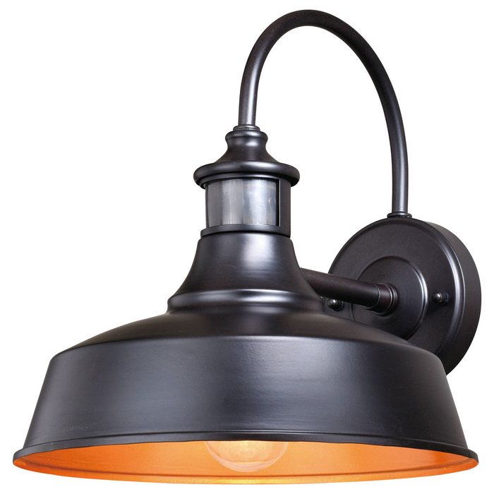 A black, industrial-style wall lamp illuminates a room, featuring a dome shade with an orange interior and a mounted sensor above the light source.
