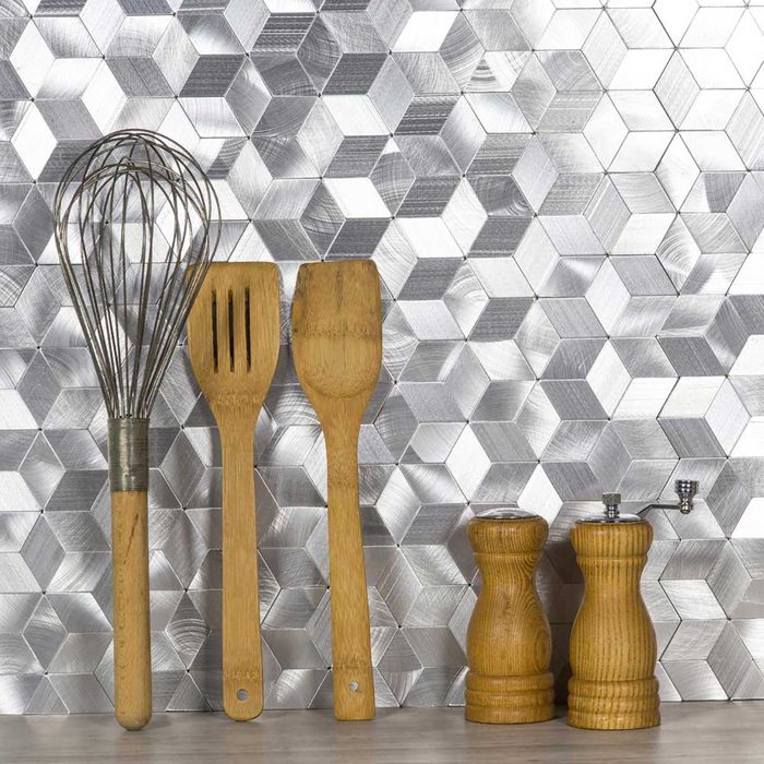 A whisk, wooden spatula, turner, and salt and pepper shakers rest against a silver hexagonal-patterned background, suggesting a kitchen setting.