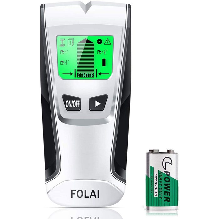 A digital stud finder displays a green screen with measurement indicators while resting beside a battery, suggesting it's ready for use in locating studs.