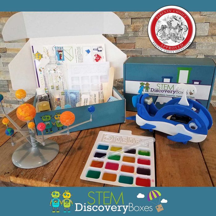 A colorful STEM discovery box displays a solar system model, educational materials, and a paint palette on a wooden surface against a stone backdrop.