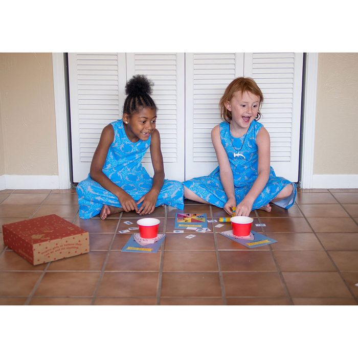 Two children in blue dresses sit on tiled flooring, playing a game with cards and red cups, looking happy in a cozy indoor setting.