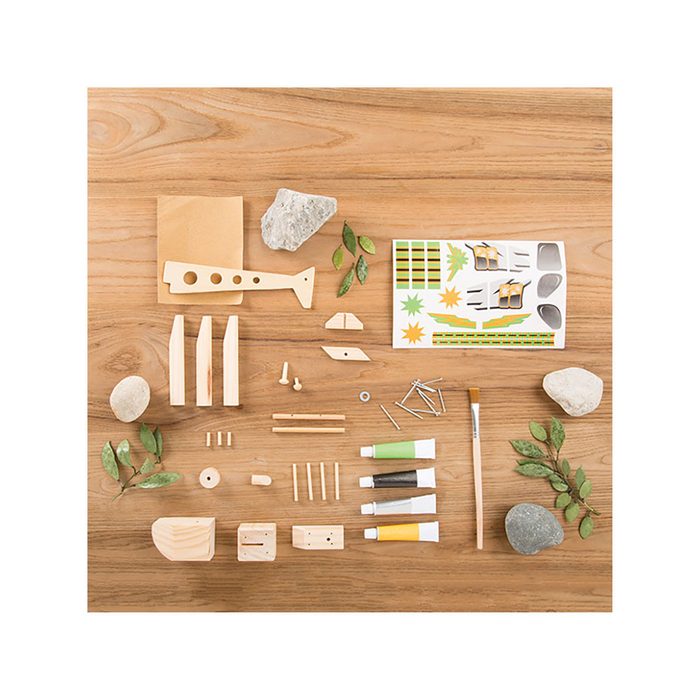 Wooden craft parts and tools are arranged on a wooden surface, including paint tubes, stickers, rocks, and leaves, suggesting a creative project setup.