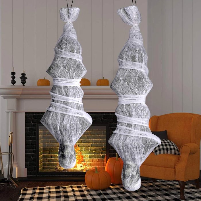 Two ghostly figures wrapped in white webbing hang from the ceiling, with an orange armchair and a fireplace adorned with pumpkins in the warm indoor setting.