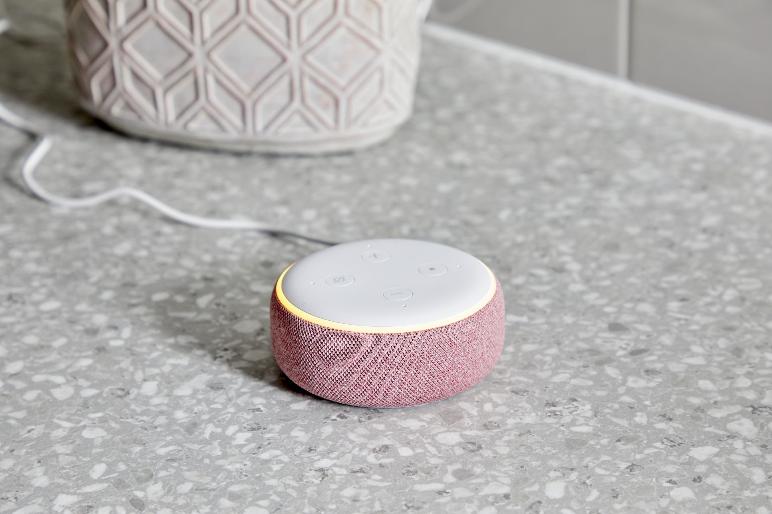 A circular, fabric-covered smart speaker rests on a speckled countertop, plugged in, with illuminated control buttons on top.