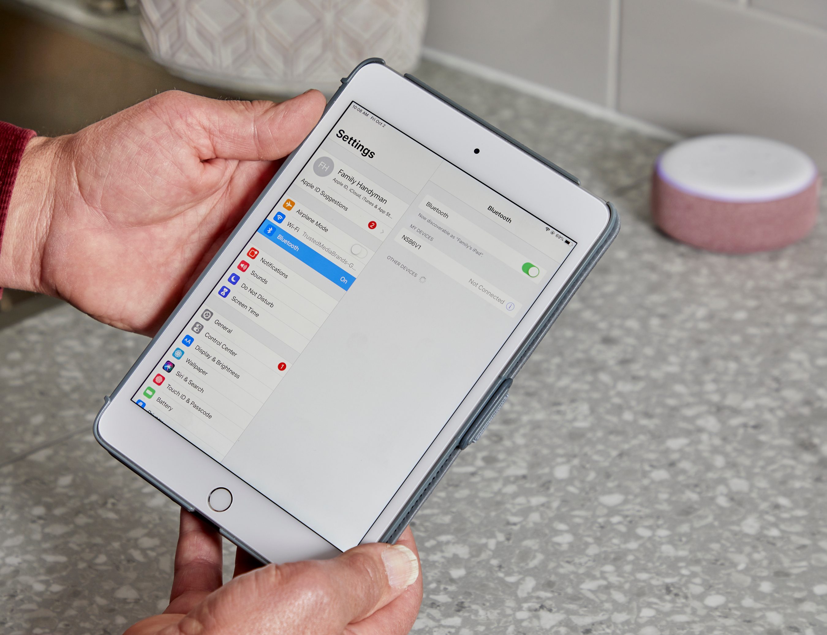 A hand holds a tablet displaying Bluetooth settings. A smart speaker sits nearby on a countertop, suggesting a tech-friendly kitchen environment.