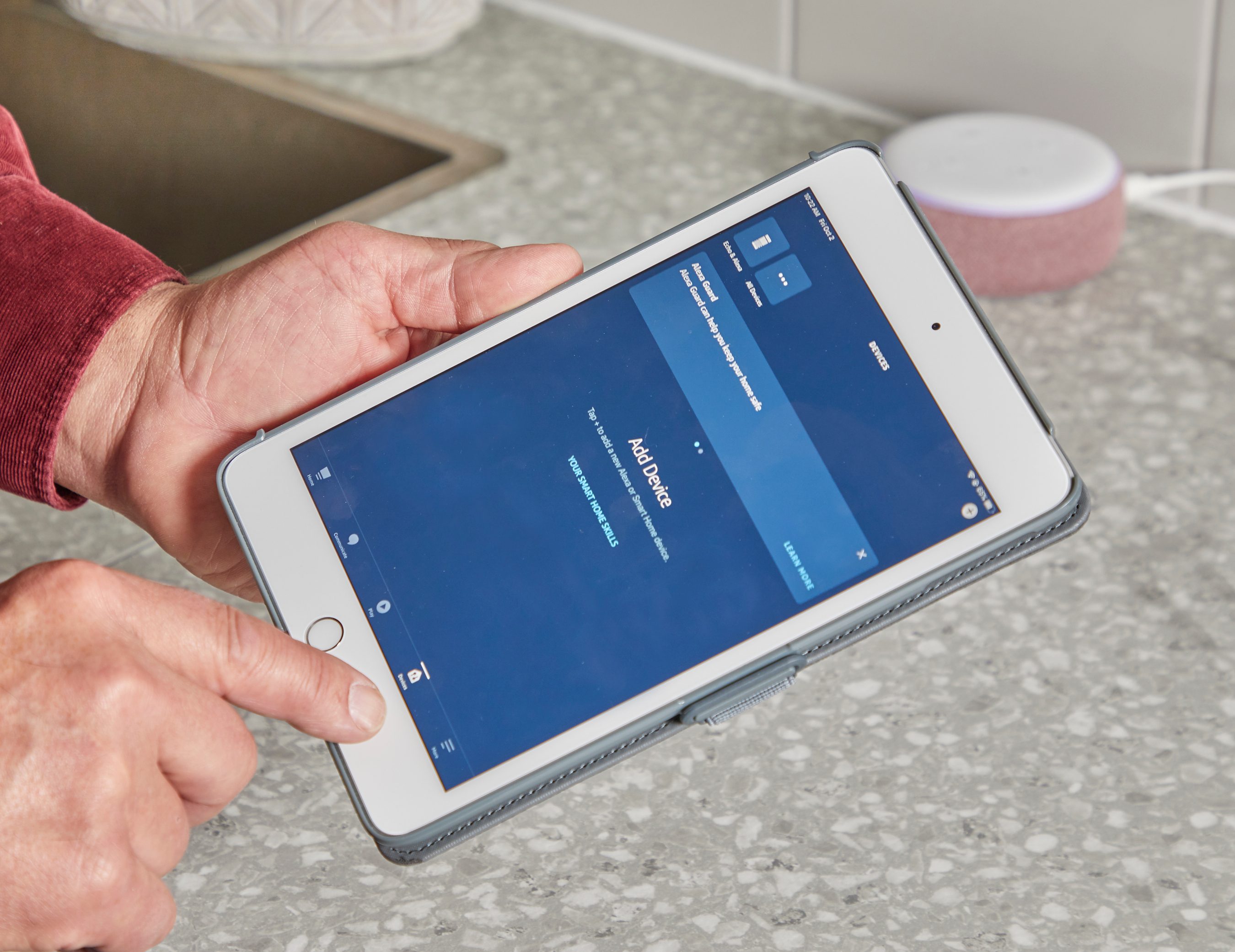 A hand holds a tablet displaying a blue screen with options. It's set on a textured countertop near a small device.