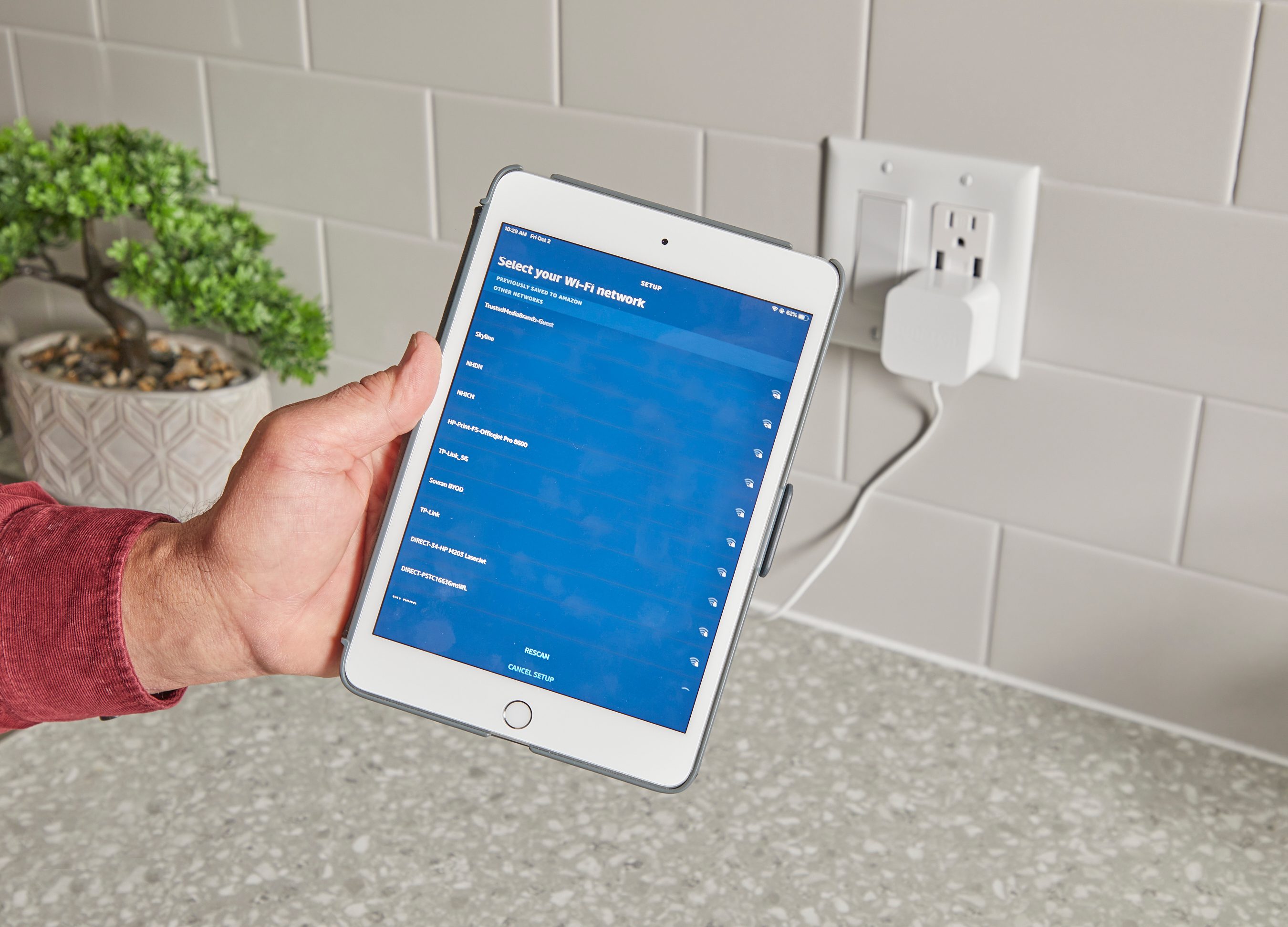 A hand holds a tablet displaying Wi-Fi network options, near a wall outlet with a plug. The setting features a textured countertop and a potted plant.