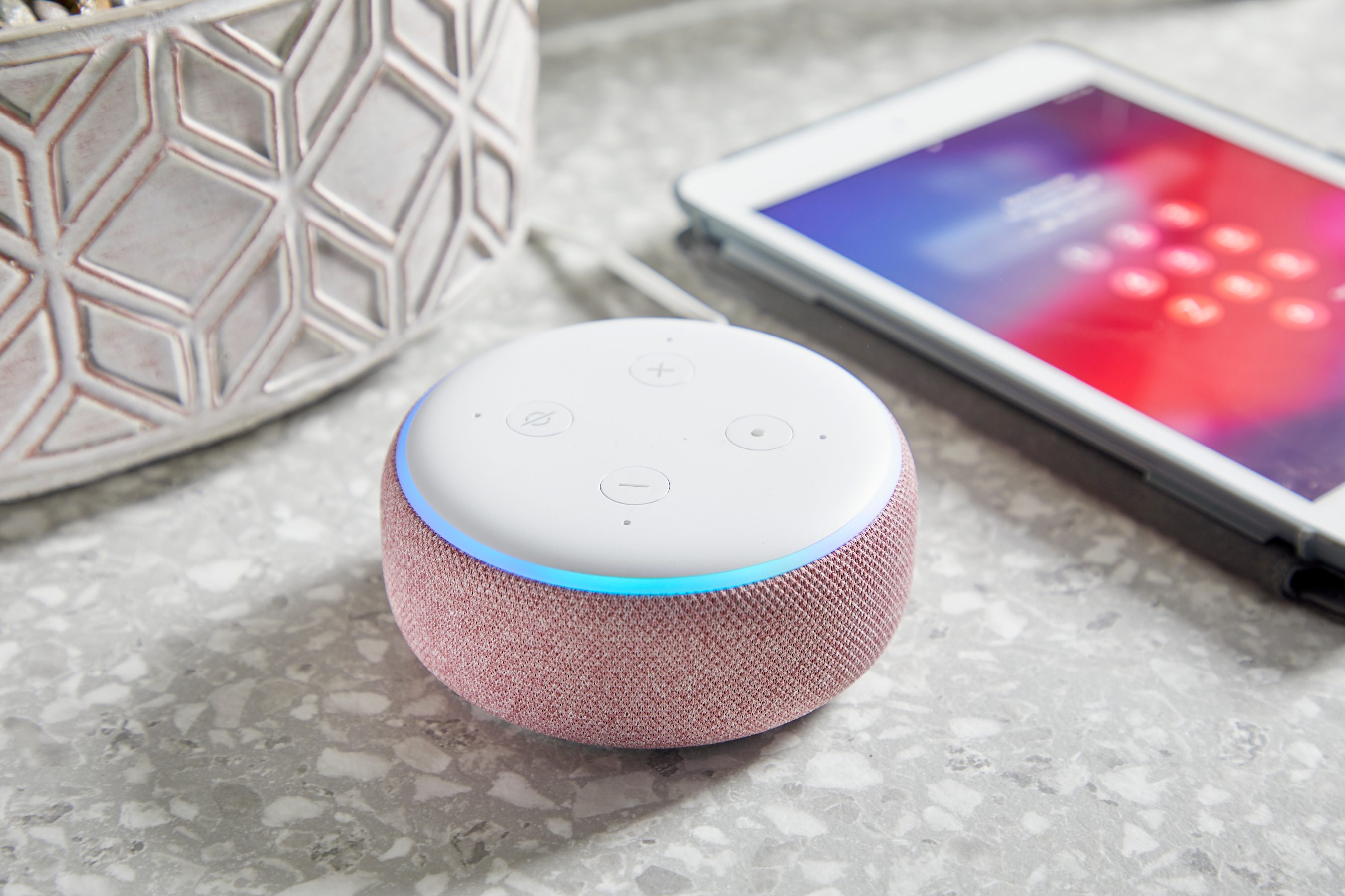 A smart speaker sits on a countertop, glowing softly, beside a tablet displaying a locked screen. A patterned planter is nearby, enhancing the setting.