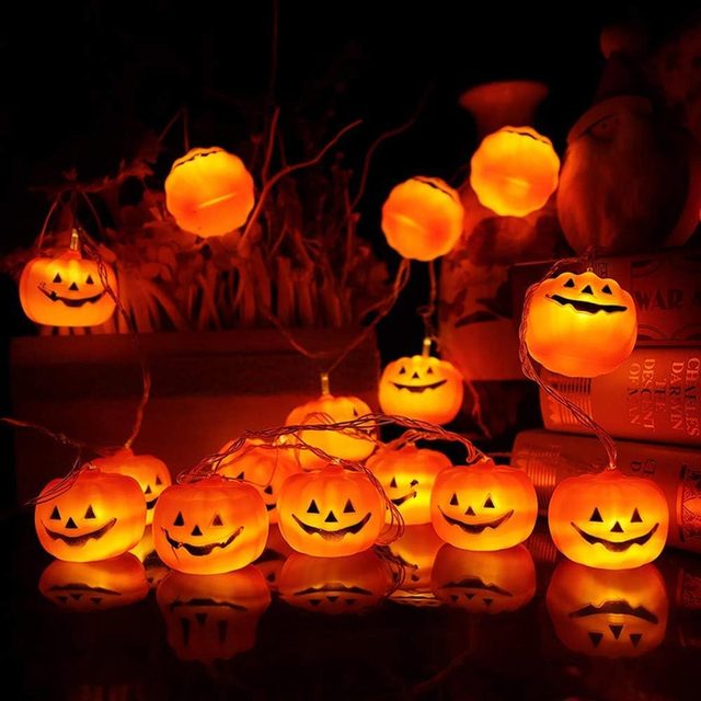 Glowing orange pumpkin-shaped lights are arranged among decorative plants and books, creating a festive, spooky atmosphere in a dark setting.