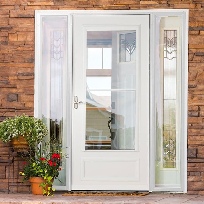A white door stands open, revealing a bright interior, flanked by glass panels featuring decorative designs, with potted flowers at the base on a stone wall.