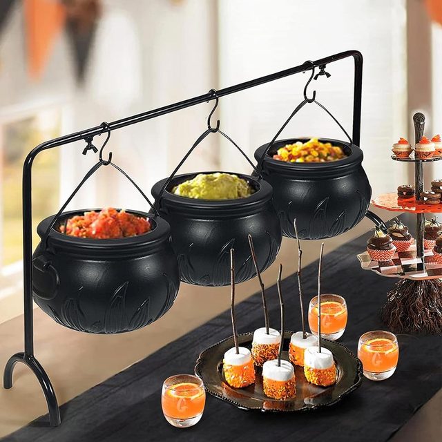 Three black cauldrons hang from a metal stand, containing colorful dishes. A dessert platter and glasses of a colorful beverage sit on the table nearby.