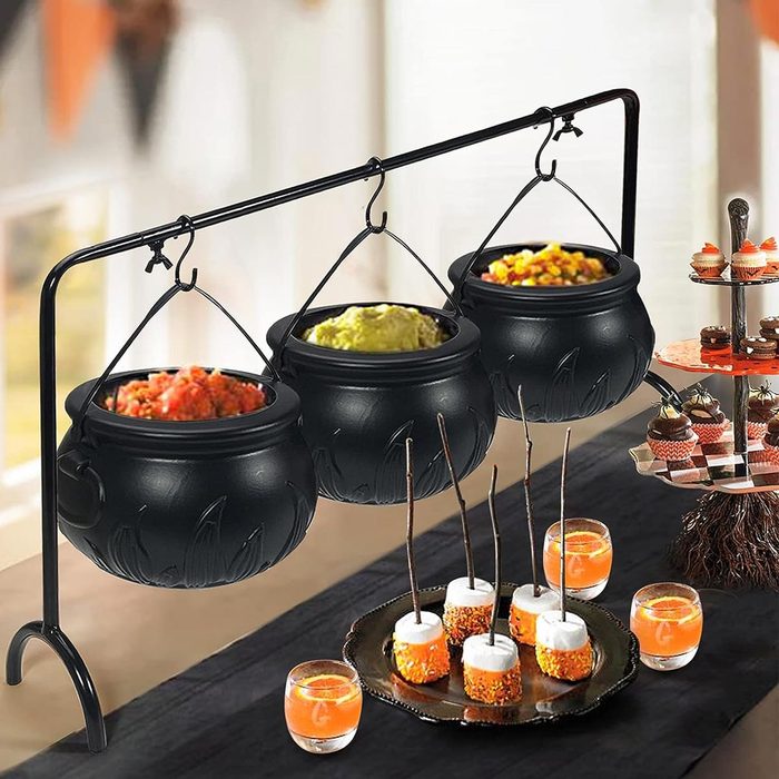 Three black cauldrons hang from a metal stand, containing colorful dishes. A dessert platter and glasses of a colorful beverage sit on the table nearby.