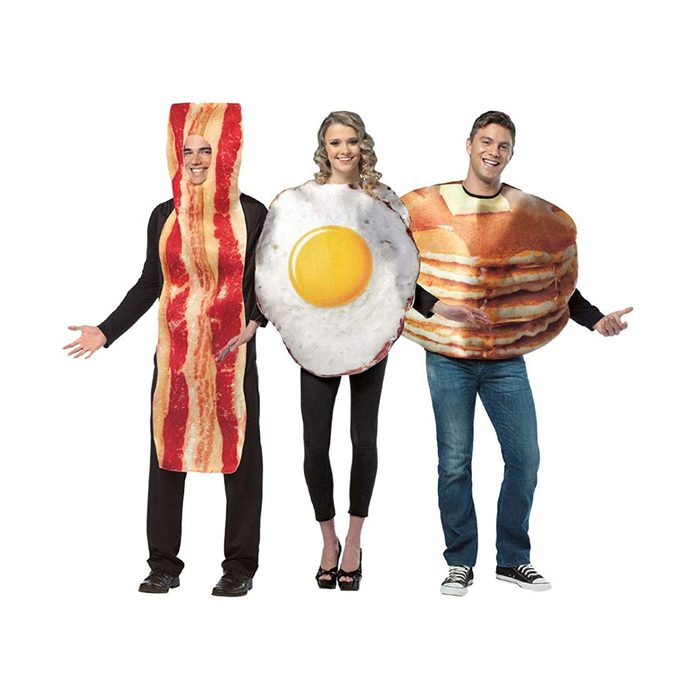 Three people wear large, food-themed costumes representing bacon, an egg, and pancakes, standing together and smiling against a plain background.