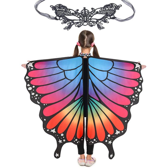 A child wears a vibrant butterfly wing costume, spreading the colorful wings wide while standing still, with a decorative mask positioned above.